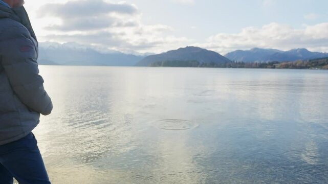 Man skipping stones on lake waters in winter season in New Zealand. Young man enjoys throwing rocks and skipping them on lake in Wanaka South Island during road trip or winter vacation outdoors