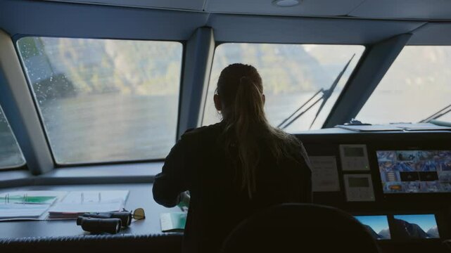 Female ship captain standing in vessel bridge looking through binoculars. Passing through mountains and fiords woman in charge. Big windows to outside as boat sails at sea or lake. Rear view back shot