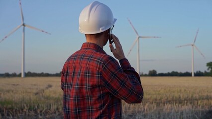 Man engineer, wearing a white protective helmet is talking by smartphone in a field with wind turbines, as the sun sets. Clean energy and engineering concept