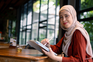 A confident and cheerful woman wearing a hijab is seated at a cafe table, engaging with a tablet....