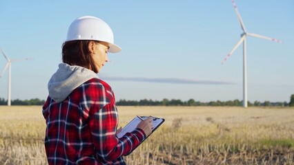 Woman engineer wearing a white protective helmet is taking notes with a clipboard in a field with wind turbines, as the sun sets. Clean energy and engineering concept