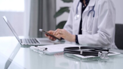 Fototapeta premium Stethoscope and clipboard are lying on the glass table while doctor woman is using a smartphone at the background. Medicine concept
