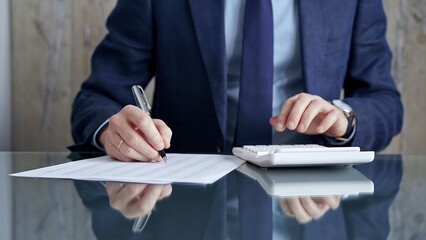 Businessman or auditor analyzing financial documents with magnifying glass and calculator at his glass office desk in formal blue jacket. Business people concept