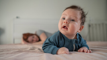 A three-month-old boy lies on his stomach on the bed and his mother sleeps behind him. Postpartum...