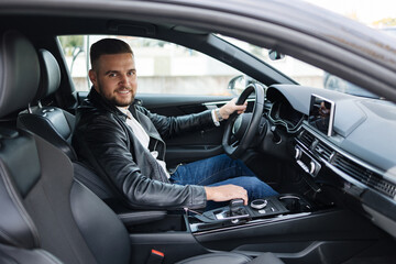 Success in motion. Handsome young man in leather jacket smiling while driving a car. Side view from inside