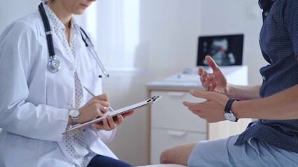 Doctor woman is taking notes on a clipboard while a male patient describes his symptoms in clinic...