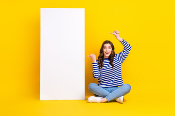 Full length photo of astonished cheerful lady sit floor indicate fingers empty space banner isolated on yellow color background