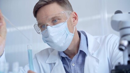 Fototapeta premium Man with safety glasses and blue mask is working with lab tubes using a pipette in a modern laboratory, close-up of researcher. Science and medicine