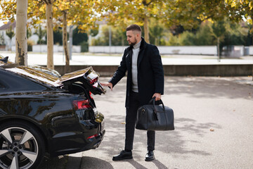 Businessman takes a bag and closes trunk of his luxury car and go to airport. Stylish bearded man in coat on auto parking