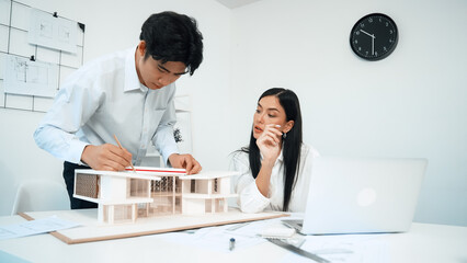 Professional male asian architect using ruler to measure house model length while young beautiful caucasian colleague using laptop to analyzed data on meeting table with house model. Immaculate.