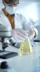Fototapeta premium Man scientist researcher, wearing white protective gloves, mask and glasses, is holding a yellow chemical solution inside Erlenmeyer flask in a laboratory, vertical portrait view. Science and medicine