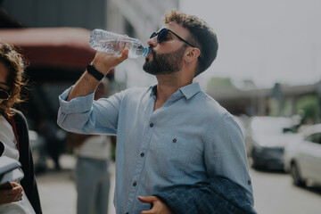 A casually dressed man drinking water from a bottle while standing outside on a sunny day. He wears...