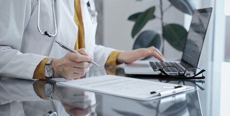 Fototapeta premium Doctor woman is writing on a document on a glass desk, with a laptop and tablet visible. Work space concept in Medicine