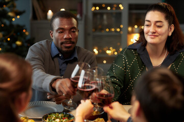 Medium shot of smiling Middle Eastern woman and Black man clinking glasses of red wine celebrating Christmas with friends during festive dinner at home party