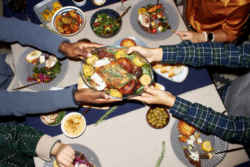 Top view of unrecognizable man passing homemade baked chicken to friend at dining table served with variety of festive dishes during Christmas dinner, camera flash, copy space