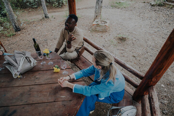 A group of friends enjoying a relaxing day outdoors, seated at a wooden table surrounded by nature. They are engaged in conversation, sharing a bottle of wine in a forest setting.