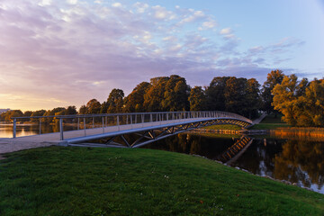 A modern pedestrian bridge stretching over a calm river, surrounded by lush greenery and trees. The bridge has a sleek, minimalist design. Pastel hues, indicating either sunrise or sunset. 