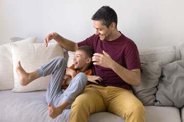 Happy Hispanic dad and joyful son kid playing tickling game on home sofa, laughing, sitting close,...