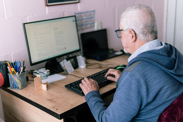 Rear view of an elderly man sitting at a desk, using a computer in a home office setting