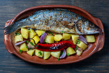 Delicious baked big sea bass fish with spices in a clay plate and fried vegetables, closeup, top view. Cooked sea bass with potatoes, red chili pepper, onion and garlic