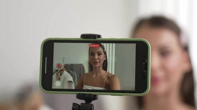 Young social medial content creator woman make fashion video. Blogger smiles to camera using selfie stick and light ring while making persuasive online clothing sell vlog to audience or follower. Adit