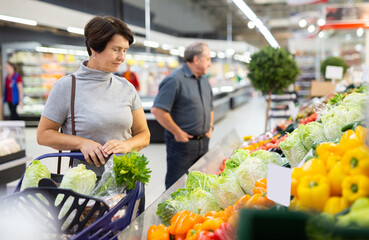 Elderly woman examines china cabbage in vegetable section to choose juiciest cabbage