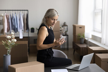 Focused aged woman warehouse employee retail client manager contact customer by social media networking with supplier by phone via mobile internet. Elderly lady web shop worker confirm order by email