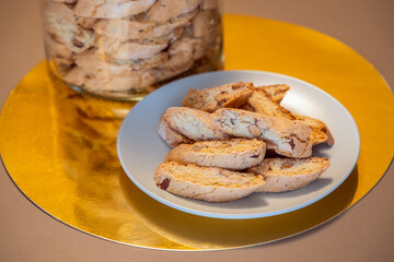 Plate of almond cantucci sits on a reflective gold surface next to a glass jar filled with more cookies. The vibrant background highlights the cookies crunchy texture and nutty flavor