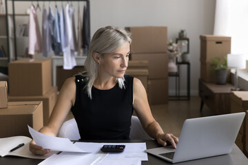 Aged woman web shop owner use pc calculator in accounting work pay bills online via internet app. Focused entrepreneur calculate price of product on web marketplace based on delivery cost and expenses