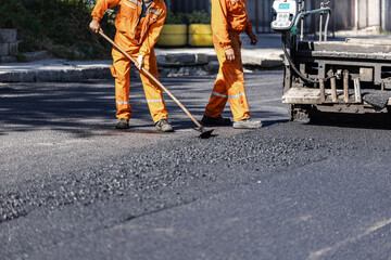 Two construction workers in orange uniforms laying asphalt on a road during daylight at a...