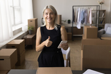 Excited senior woman postal worker mail service manager hold cellphone on workplace look at camera with thumb up gesture. Internet shop keeper promote perfect goods service approve easy fast delivery