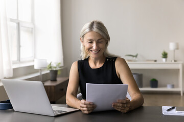 Joyful mature self employed female working from home office read paper letter with good business...