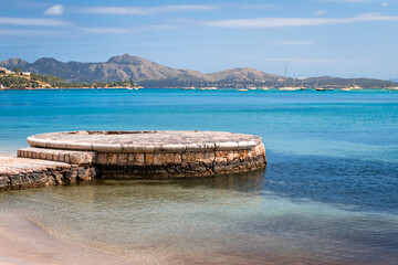 Port de Pollença beachfront in Mallorca with a circular stone pier extending into the calm, shallow waters of the bay, framed by distant Serra de Tramuntana mountains, for tourism and travel content.