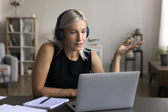 Confident mid aged female wear headset work from home office online speak on video conference webinar using virtual meeting app on laptop. Mature woman teacher give remote web lecture talk to students