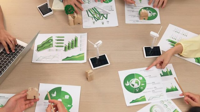 Excellent business team discussing on eco house project at meeting table with windmill design model and green power usage graph for decreasing carbon footprint, save earth. Focus on hand. Delineation.