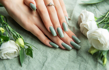Elegant hands adorned with green nail polish surrounded by white roses and greenery on a soft fabric background during daylight