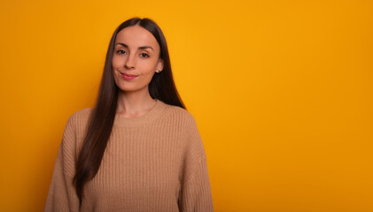 Fototapeta premium A young woman with long brown hair stands against a bright yellow background, wearing a beige sweater and smiling slightly. The image is cheerful and friendly, conveying warmth and positivity.