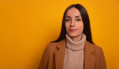 A woman in a camel coat and beige turtleneck sweater stands confidently against a yellow background. Her expression is calm and composed, exuding modern elegance and professionalism.