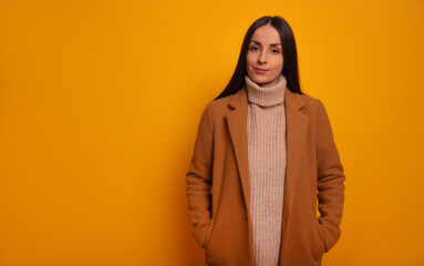 A stylish woman in a camel coat and beige turtleneck sweater stands confidently against a bright yellow background. Her expression is calm and poised, reflecting modern, elegant fashion.