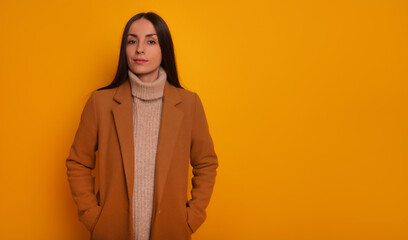 A stylish woman in a camel coat and beige turtleneck sweater gazes confidently at the camera against a bright yellow background. She exudes a calm, poised, and fashionable vibe in this portrait shot.