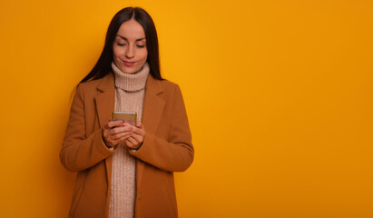A stylish woman in a camel coat and beige turtleneck sweater looks down, smiling as she uses her smartphone against a vibrant yellow background. The scene conveys a modern, tech-savvy vibe.