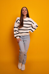 Fototapeta premium A young woman stands confidently against a bright yellow background. She wears a striped sweater, light blue jeans, and white sneakers, smiling brightly and casually posing with one hand on her hip.