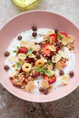 Greek yogurt with fresh strawberries, flakes and granola in a roseate bowl, vertical shot on a beige granite surface, middle closeup