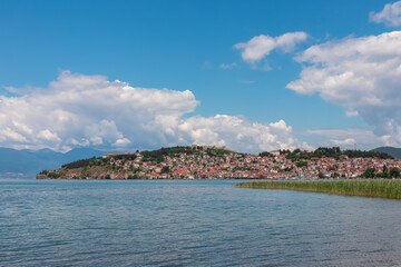 Fototapeta premium Scenic view of charming lakeside town nestled on shores of Lake Ohrid, North Macedonia. Majestic historical Samoil's Fortress, perched atop cloud covered hill. Field of reeds covering water surface