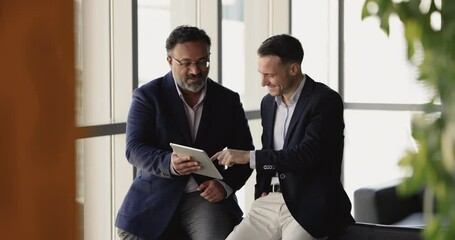 Two mature businessmen talking and laughing while using digital tablet in office, enjoy successful project completion, watch humorous video or presentation related to their work, joking at workplace