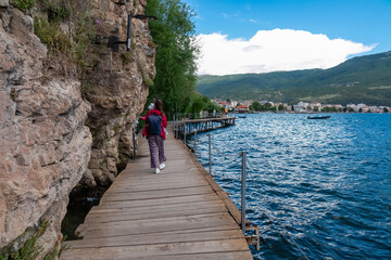 Woman with baby walking on wooden platform The Bridge of Wishes extending into calm Lake Ohrid in North Macedonia. Pier boardwalk offering scenic views of tourist lakeside town and rolling hills