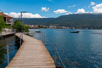 Fototapeta premium Walking on wooden platform The Bridge of Wishes extending into calm waters of Lake Ohrid in North Macedonia. Pier boardwalk offering scenic views of tourist lakeside town, rolling hills and mountains