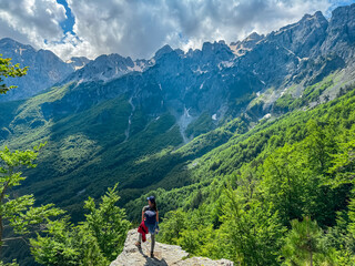 Fototapeta premium Hiker woman stands on edge of rock formation with panoramic view of Valbona Valley National park surrounded by majestic steep mountain ridges of Albanian Alps (Accursed Mountains), Albania. Wanderlust