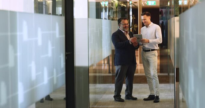 In modern office hallway two multiracial male colleagues using digital tablet, reviewing ongoing project, discuss next steps of task completion, preparing presentation for upcoming meeting. Workflow