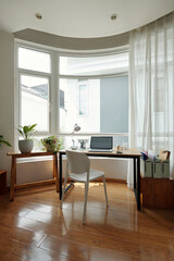 Bright home office with wooden floor, large windows, and greenery. Minimalist desk setup with computer offers serene work environment, surrounded by natural light